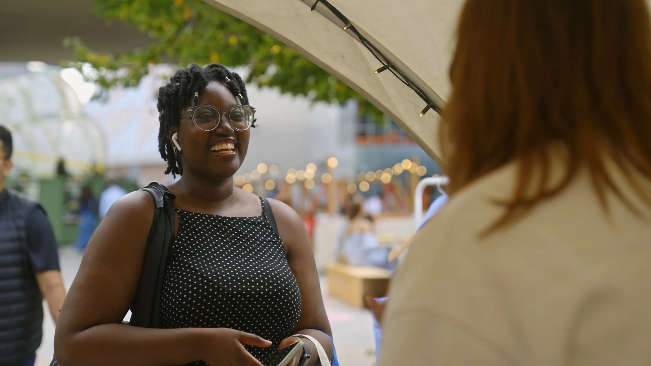 Smiling Black woman with glasses and earrings talking to a person outside at an outdoor market or fair, with string lights and trees in the background.