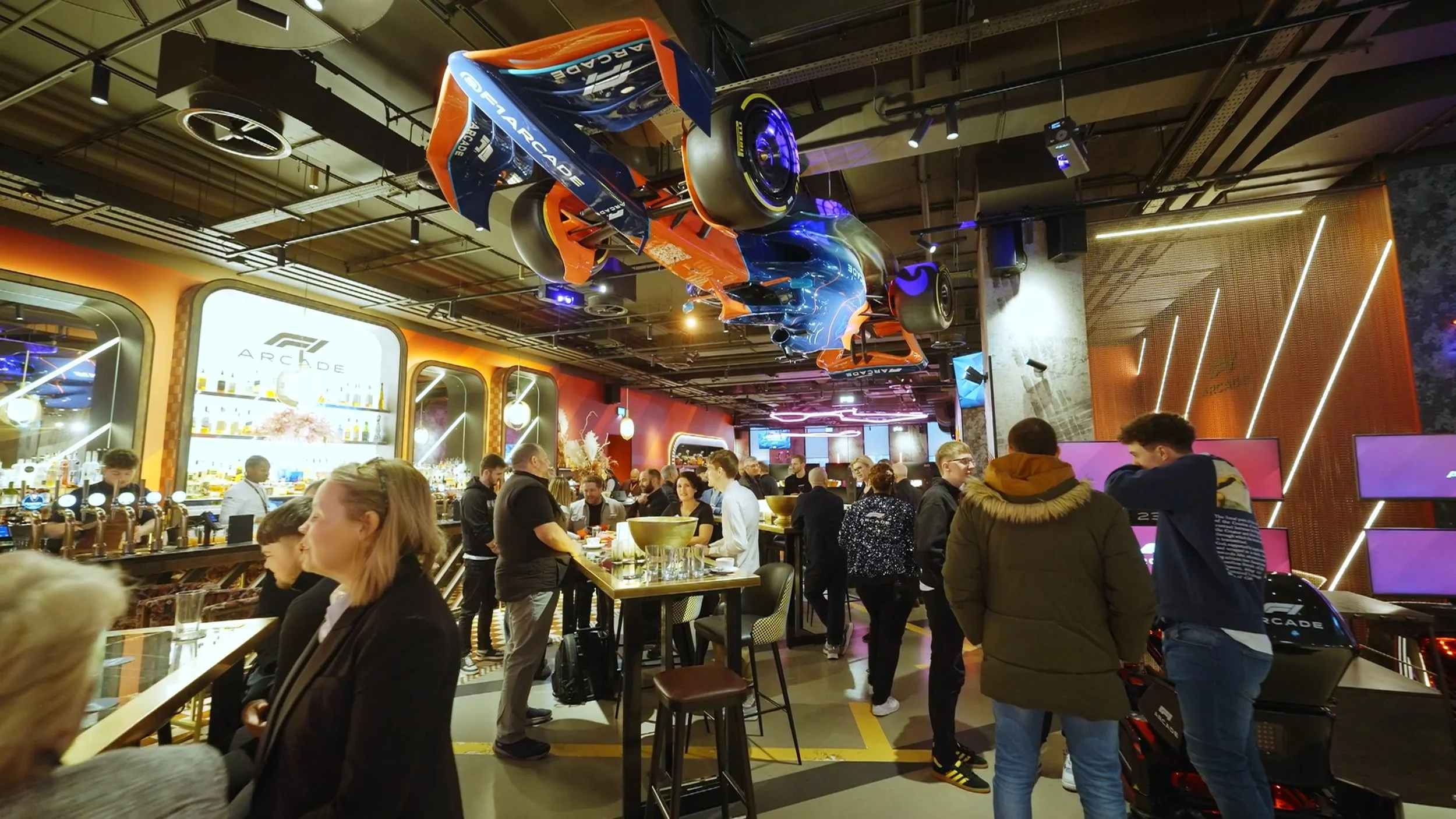 Indoor arcade bar with a Formula 1 race car hanging from the ceiling, people socializing and ordering drinks at a busy bar.