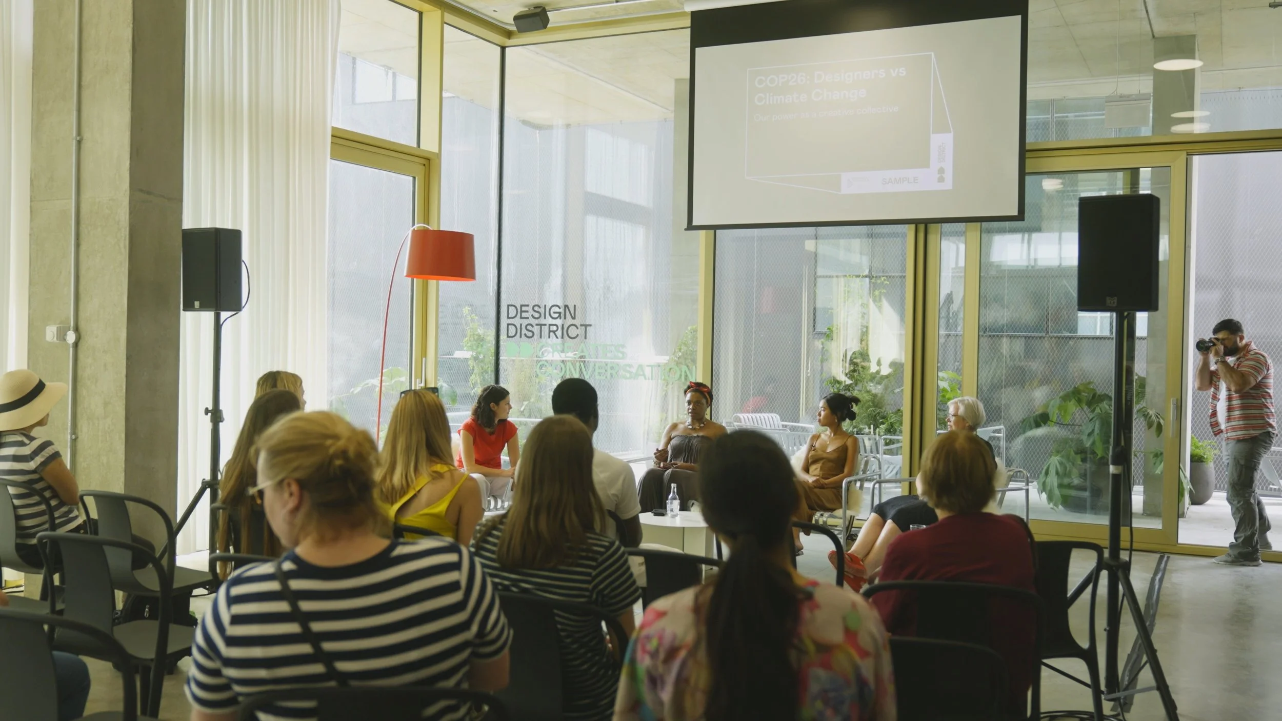 A panel of women speaking at a discussion event in a modern, bright conference room with a large screen displaying the title 'COP26: Designers vs Climate Change' and the words 'Design District' visible on the window.