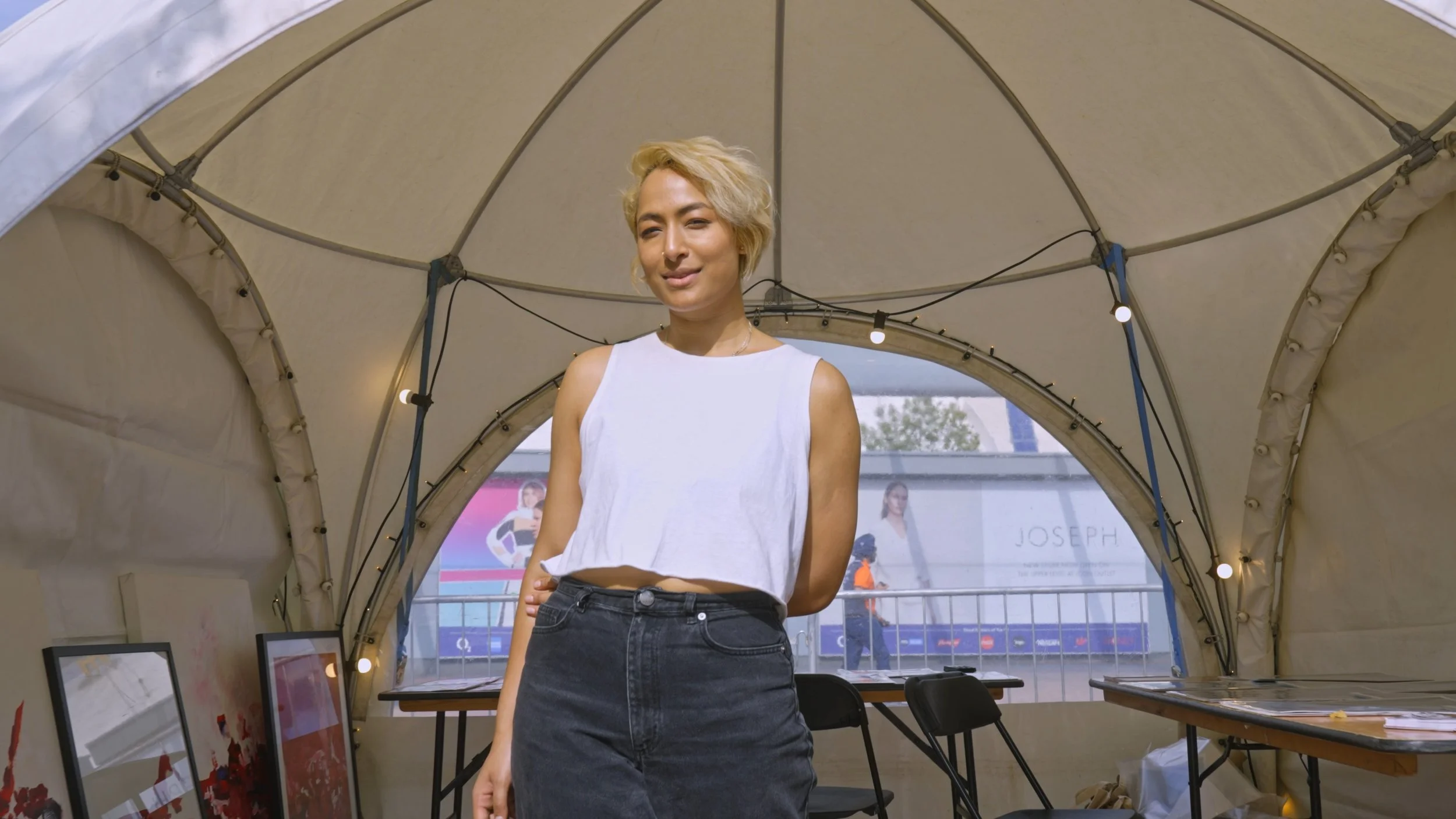 Young woman with blonde hair wearing a white sleeveless top and black jeans standing inside a beige tent, with tables and chairs behind her, and banners visible outside the tent.