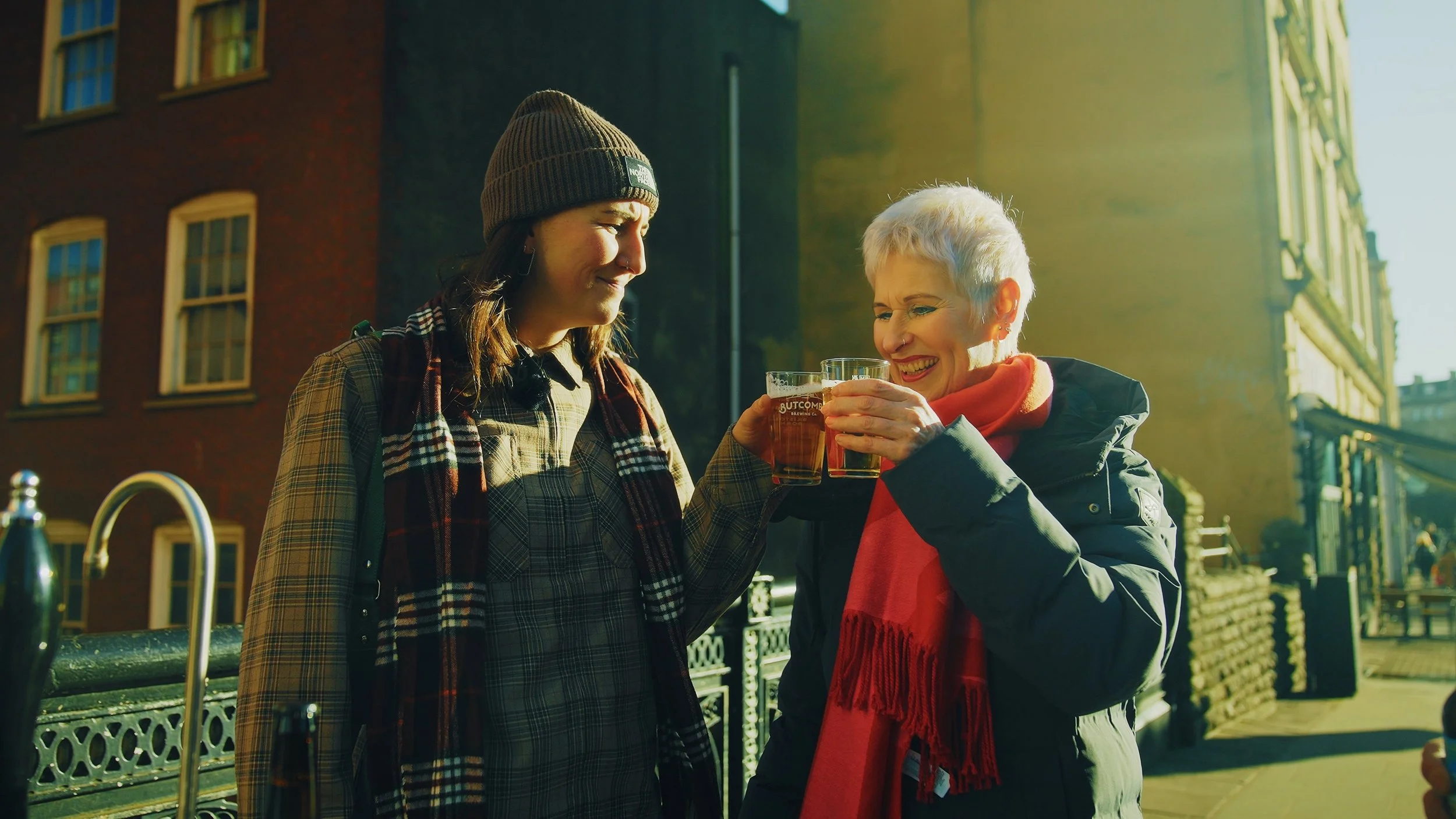 Two women clinking glasses of beer outdoors on a sunny day, smiling at each other. One woman has short white hair and a red scarf, the other has long brown hair, a beanie, and a scarf.