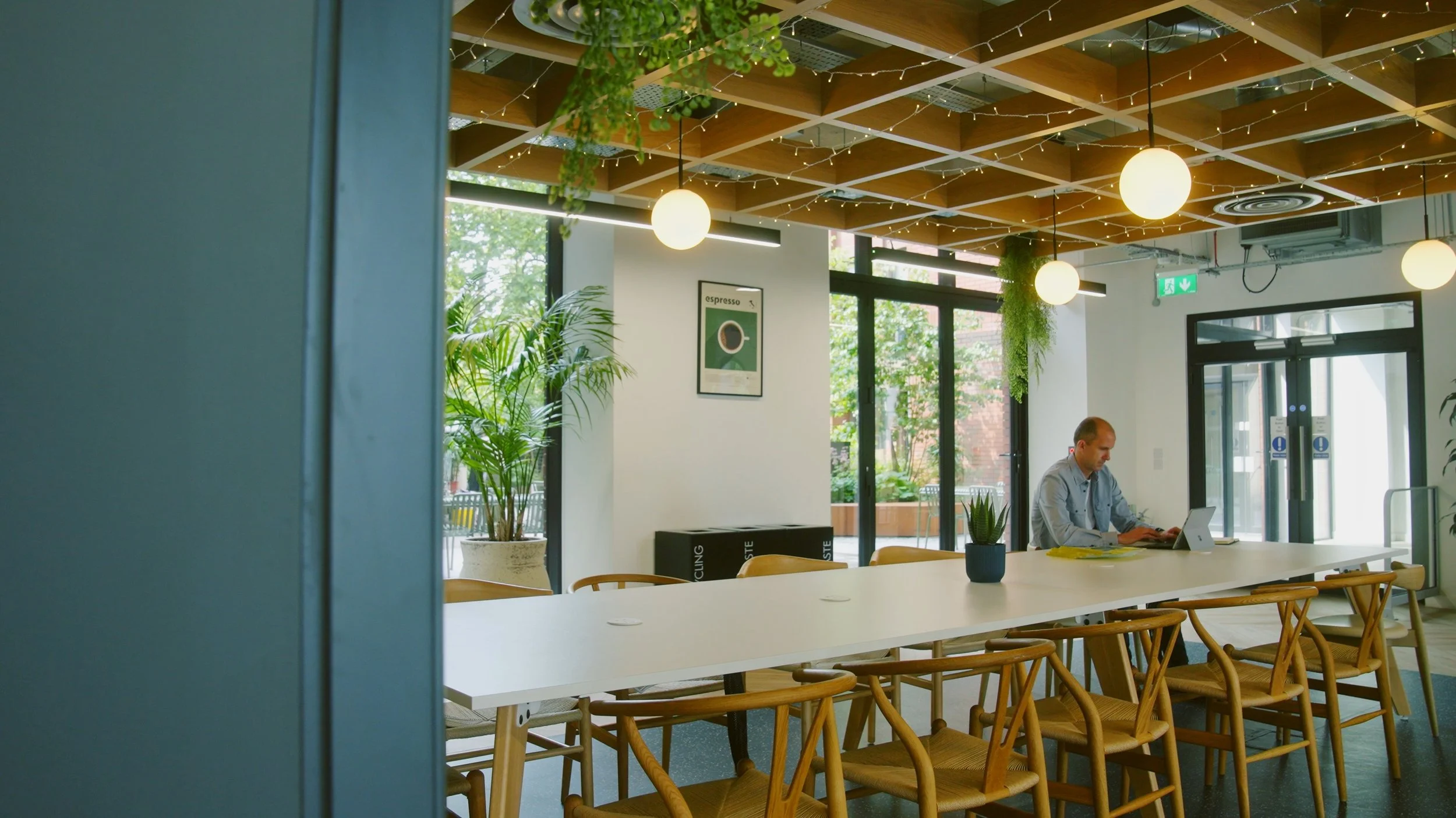A man sitting alone at a long white table in a modern, well-lit cafe with plants and hanging lights.
