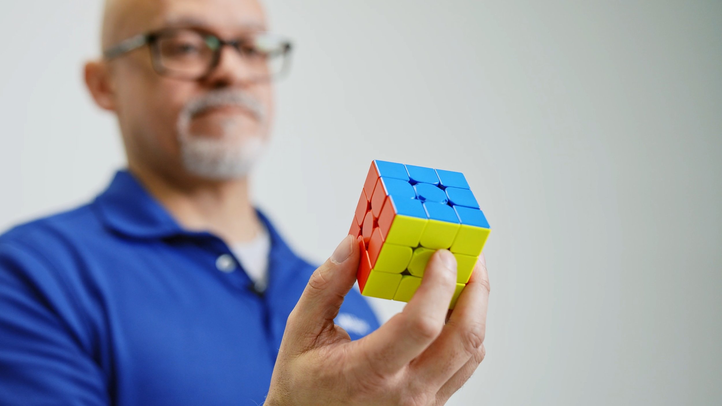A man with glasses and a gray beard holding a solved 3x3 Rubik's Cube with yellow, blue, and red faces, wearing a blue polo shirt, in front of a plain white wall.
