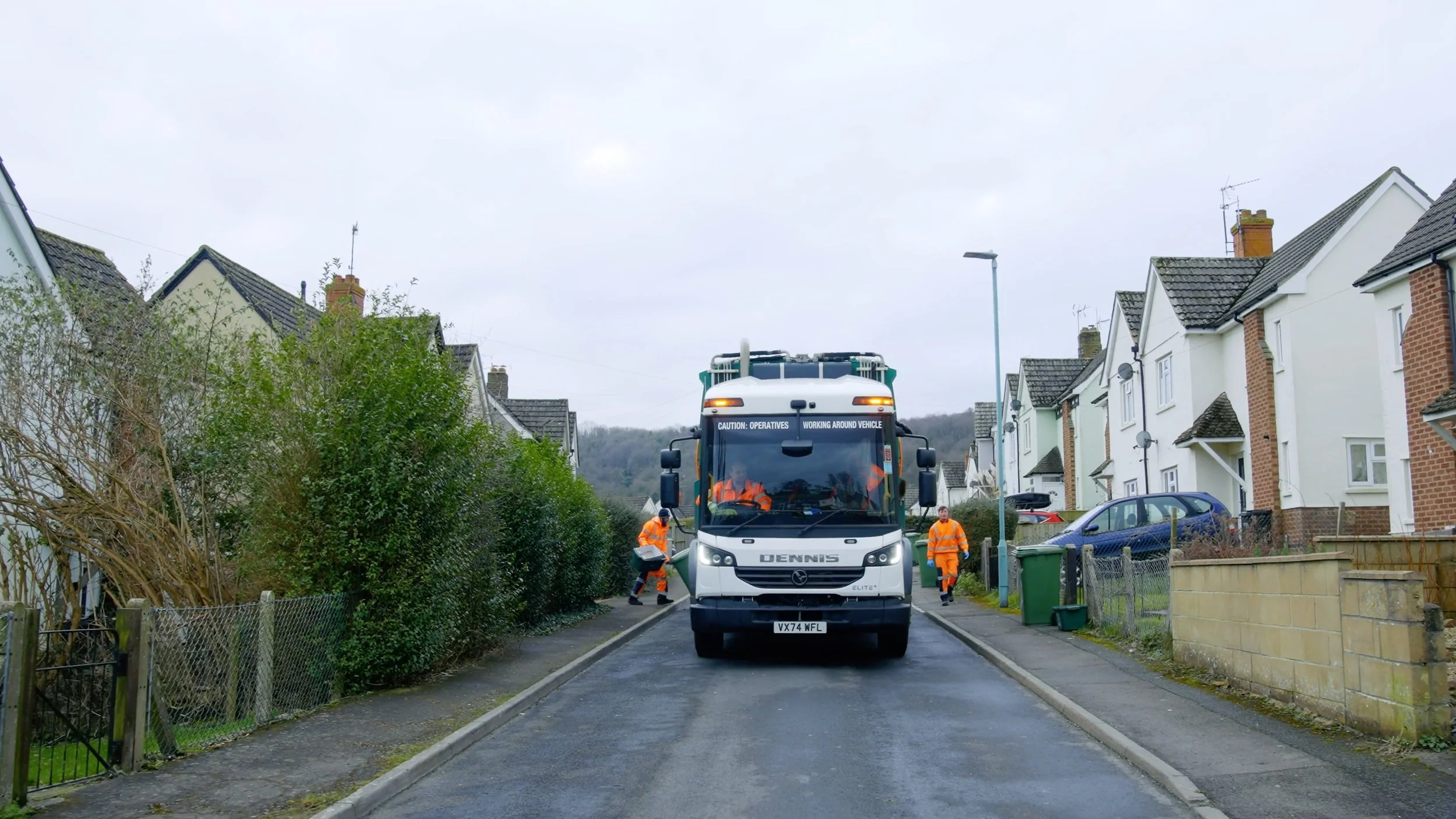Street scene with a street sweeper vehicle in the center, two workers in orange uniforms walking beside it, residential houses on both sides, and a cloudy sky overhead.