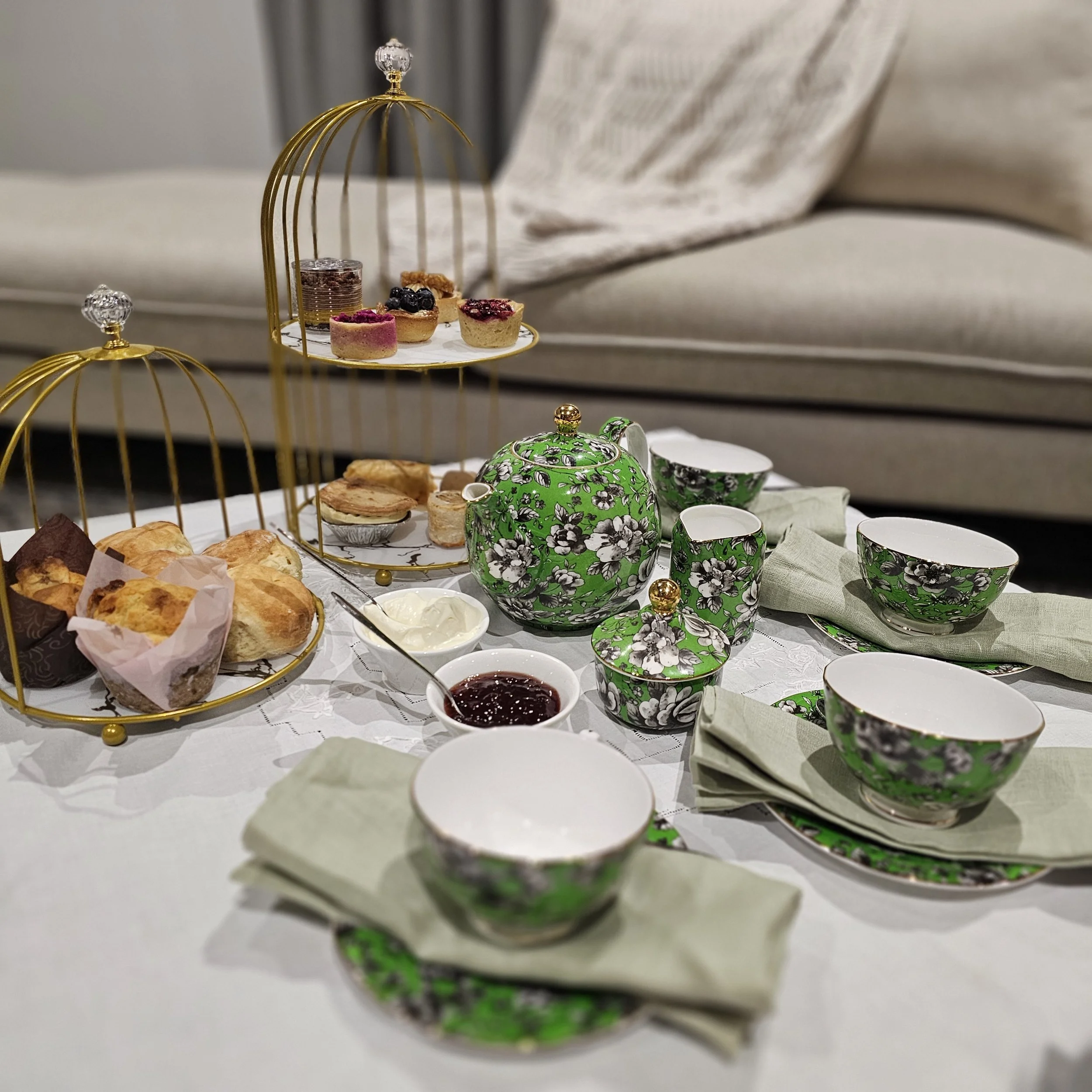 A tea set on a table with green and black floral pattern, white interior, matching cups, bowls, and serving plates, with a two-tier gold cake stand holding assorted desserts including scones, muffins, and mini cheesecakes, along with jam, cream, and additional pastries.
