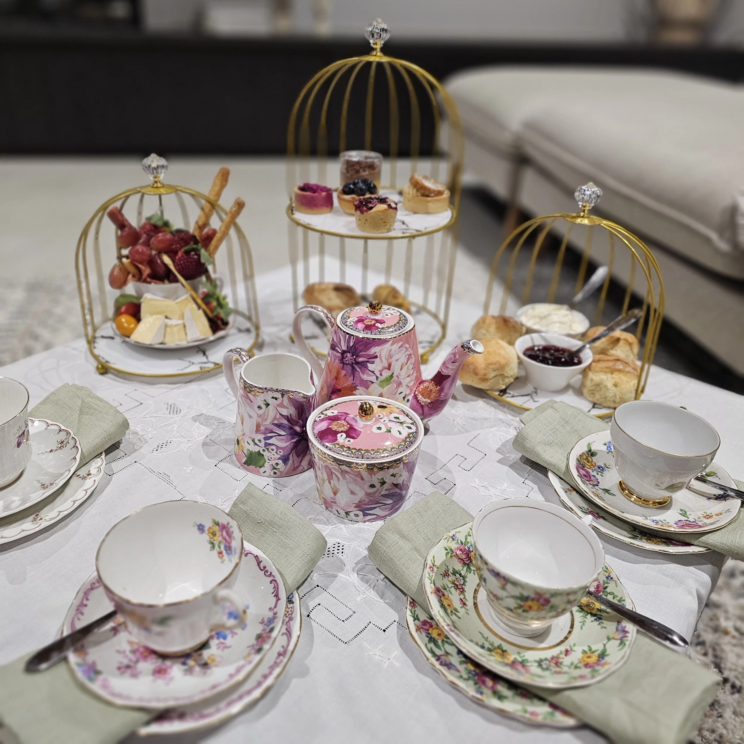 A table set for afternoon tea with floral patterned teapots, teacups, plates, and napkins. There are three gold cages holding an assortment of desserts, cheeses, fruits, and clotted cream. The table is covered with a white embroidered tablecloth.