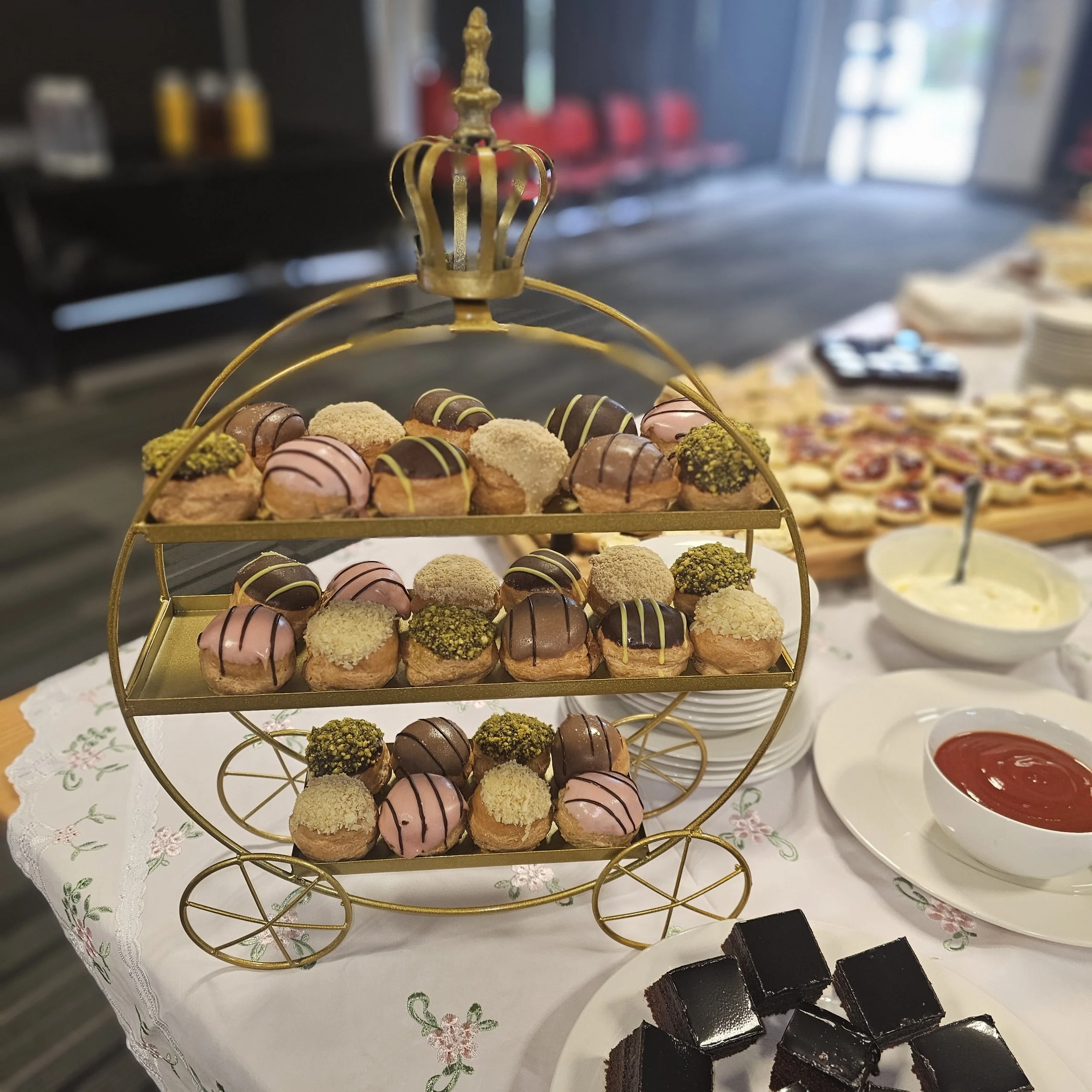 A three-tiered gold dessert tray filled with assorted small pastries topped with pink, white, and dark chocolate icing, some decorated with green pistachios, on a table with a white tablecloth. In the background, there are bowls of cream and strawberry sauce, stacked plates, and a tray of assorted cookies.