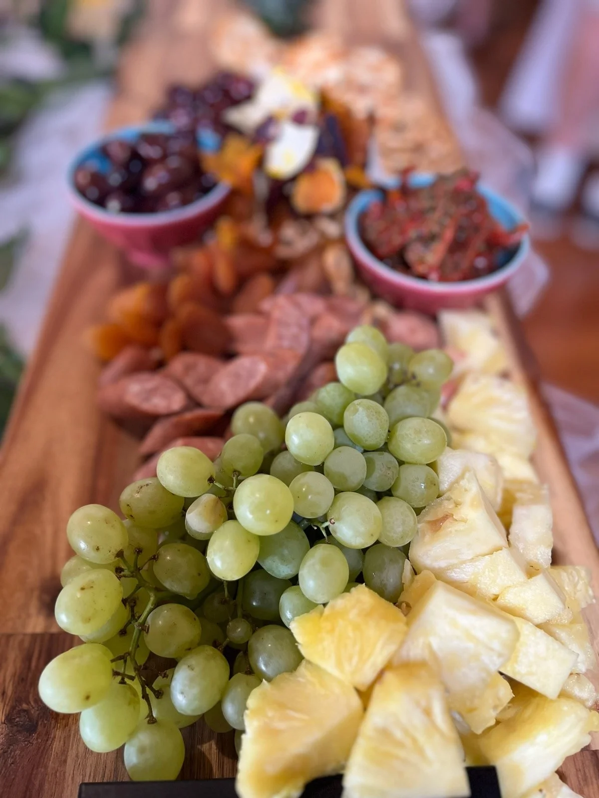 Close-up of a cheese and fruit platter with grapes and pineapple, with visible slices of cheese and small bowls of dried fruit and jam on a wooden board.