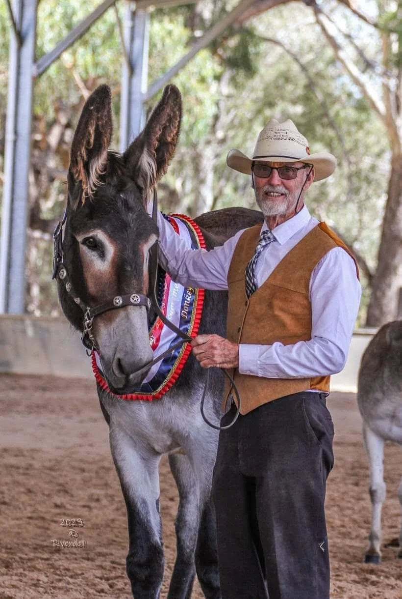 Older man in cowboy hat, glasses, white shirt, tie, and brown vest standing next to a dark brown donkey with light points and large ears, inside a covered arena with trees in the background.