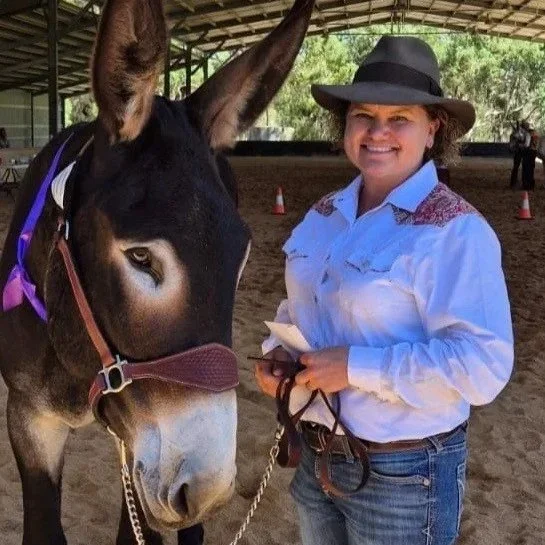 A woman wearing a hat and white western shirt standing next to a donkey in a covered outdoor area.