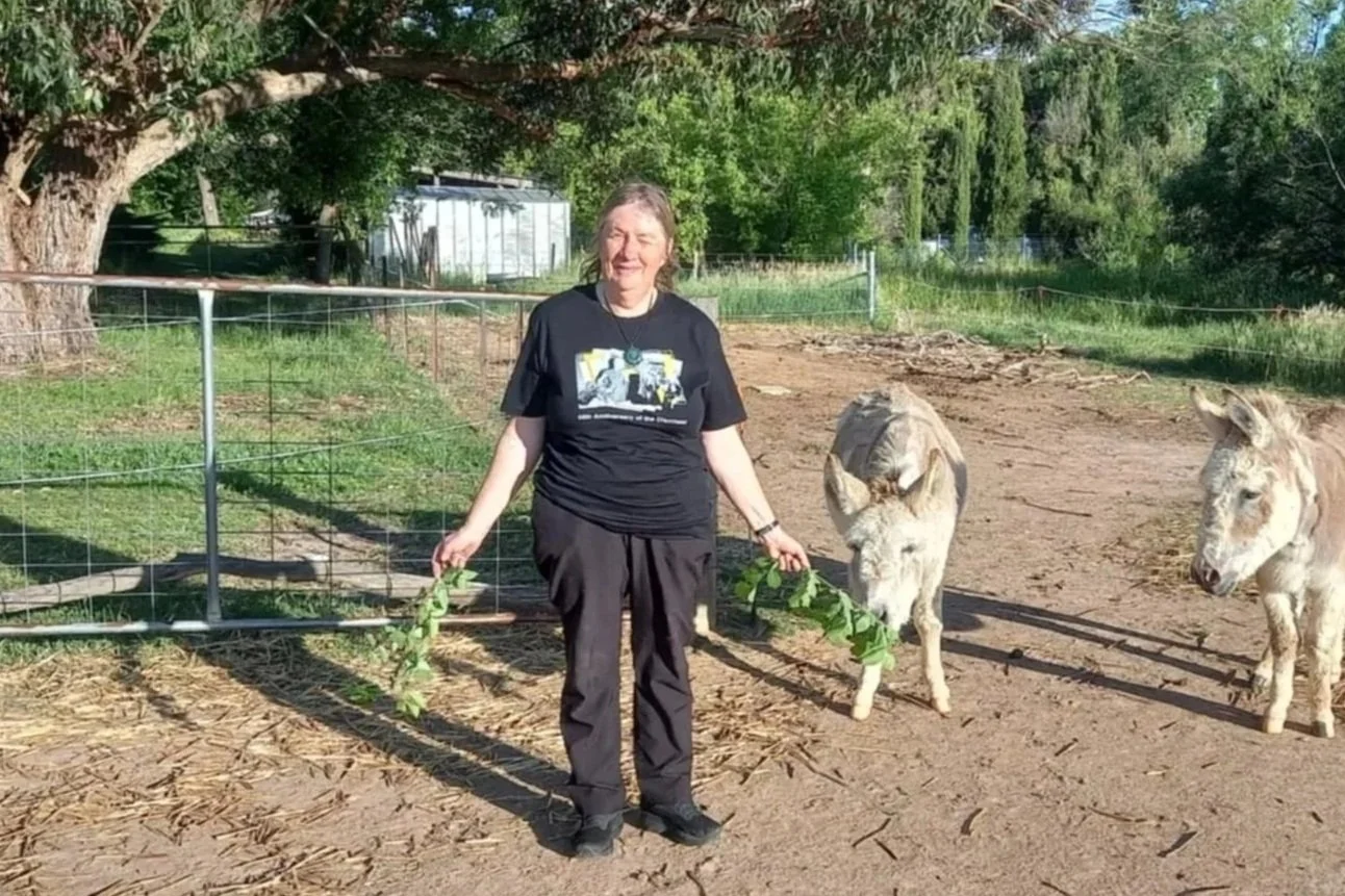 A woman standing outside on dirt ground, holding leafy branches, with two donkeys nearby. She is smiling and there are trees and a fence in the background.