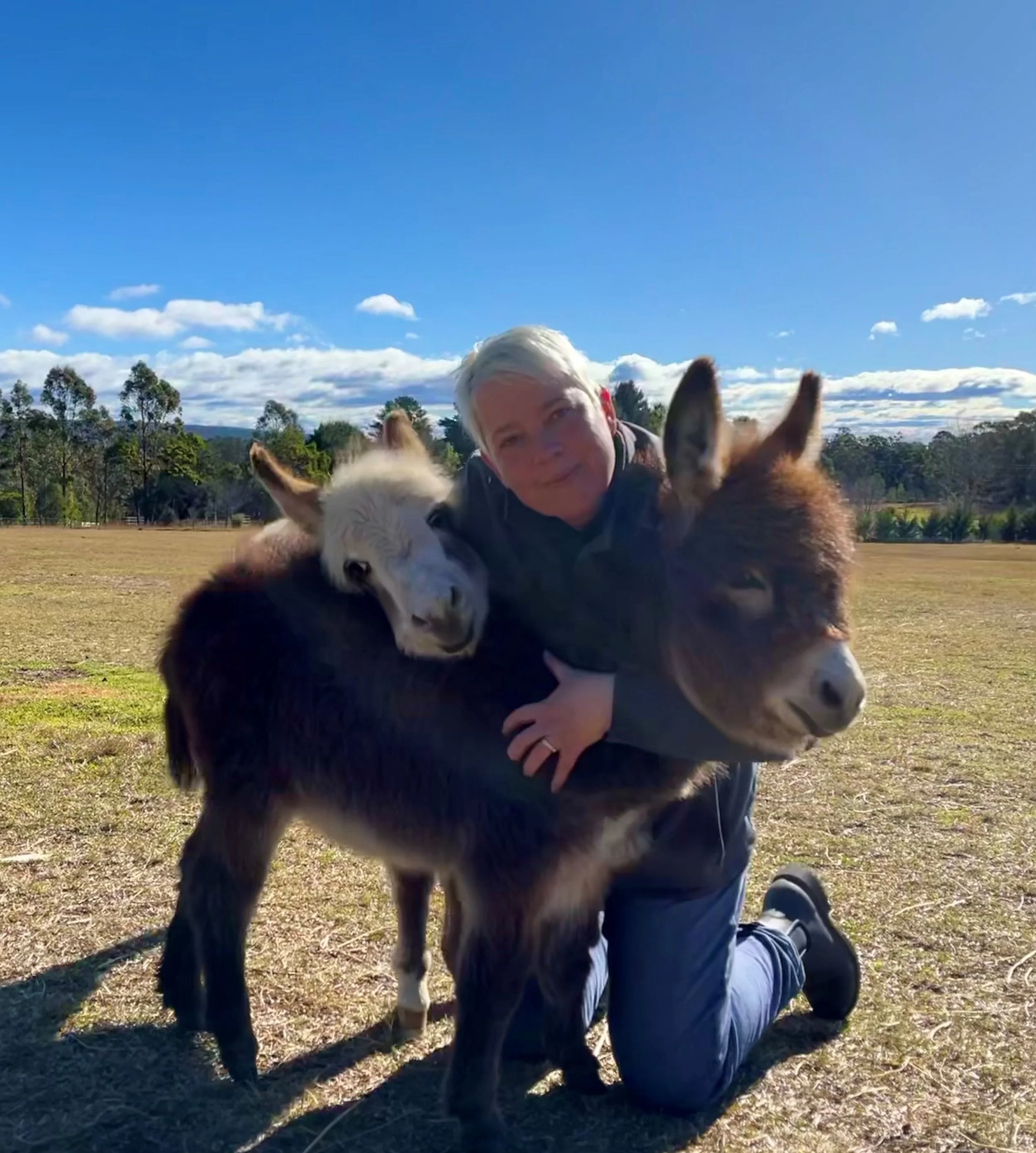A woman sitting on the grass outdoors, wearing a hat and a jacket, holding a baby donkey close to her face, as the donkey leans in to nuzzle her.