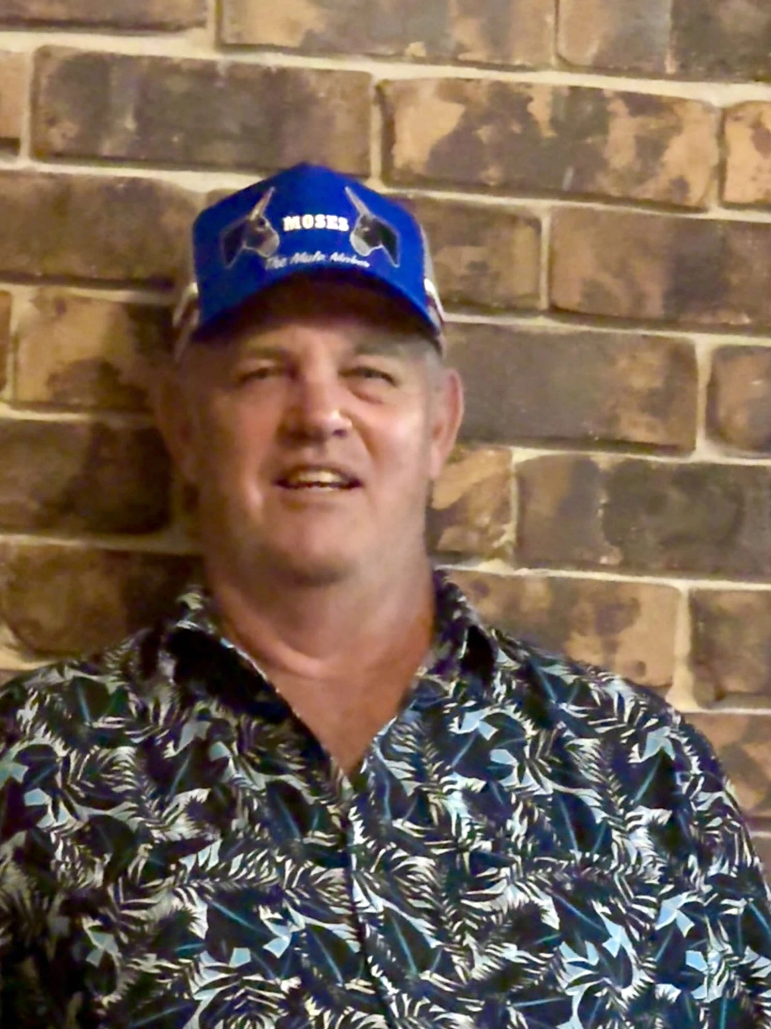 A man wearing a blue cap with the word 'MOSES' and the image of a mule on it, posing in front of a brick wall.