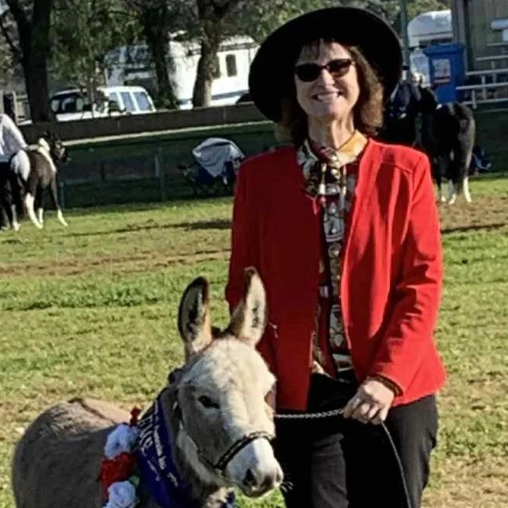 A woman wearing a black hat, sunglasses, red blazer, and patterned blouse standing outdoors with a donkey decorated with a colourful garland in a grassy area with trees and other animals in the background.