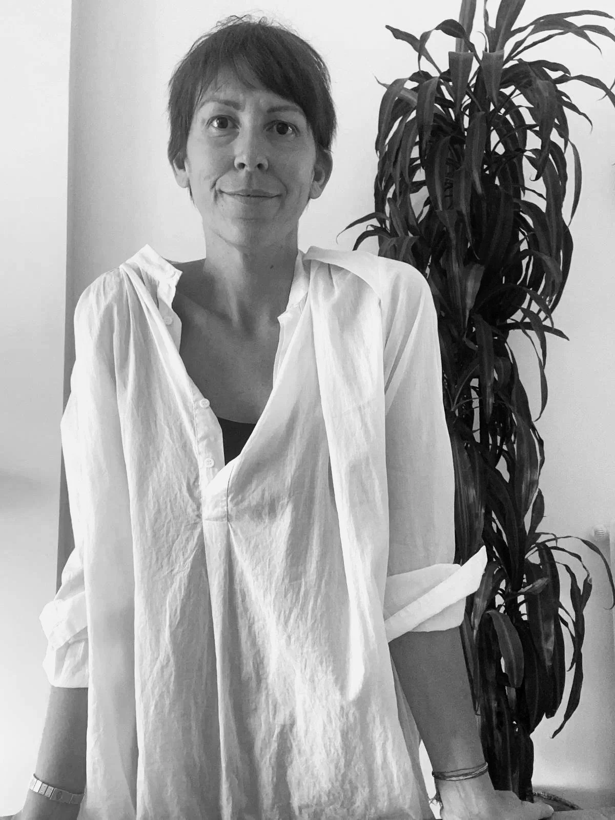 Retrato en blanco y negro de una mujer con camiseta blanca, sonriendo levemente, de cabello corto y oscuro, junto a una planta alta con hojas largas y delgadas.