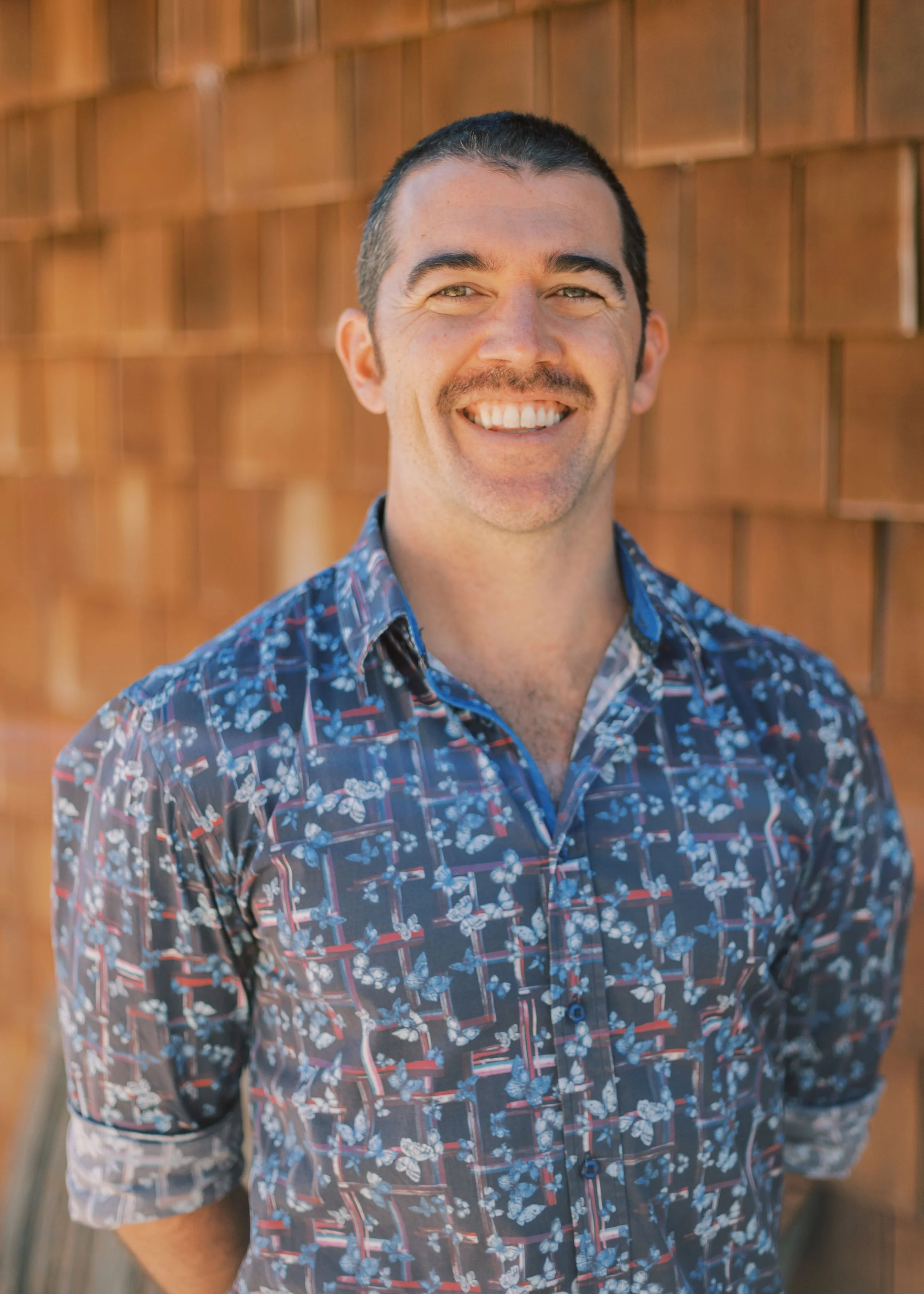 Man with patterned shirt smiling in front of wooden wall.