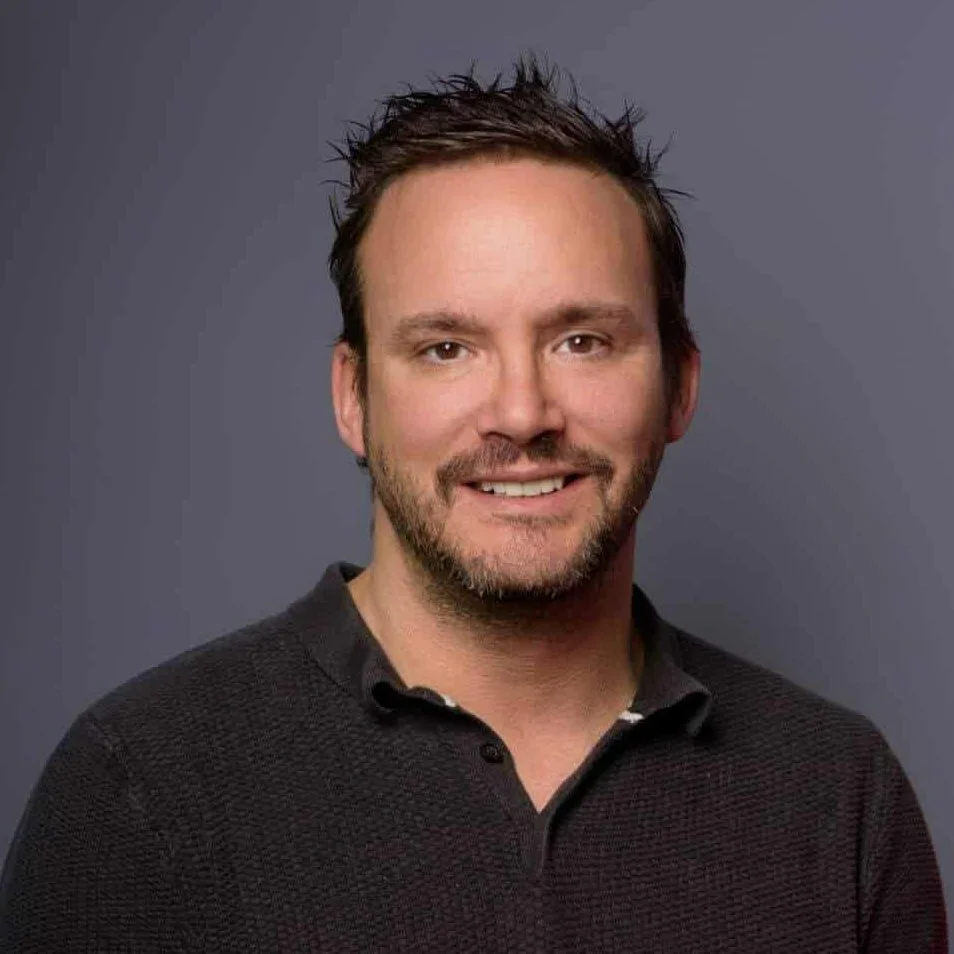 A man with short dark hair, a light beard, and a smile, wearing a dark collared shirt, standing against a plain gray background.