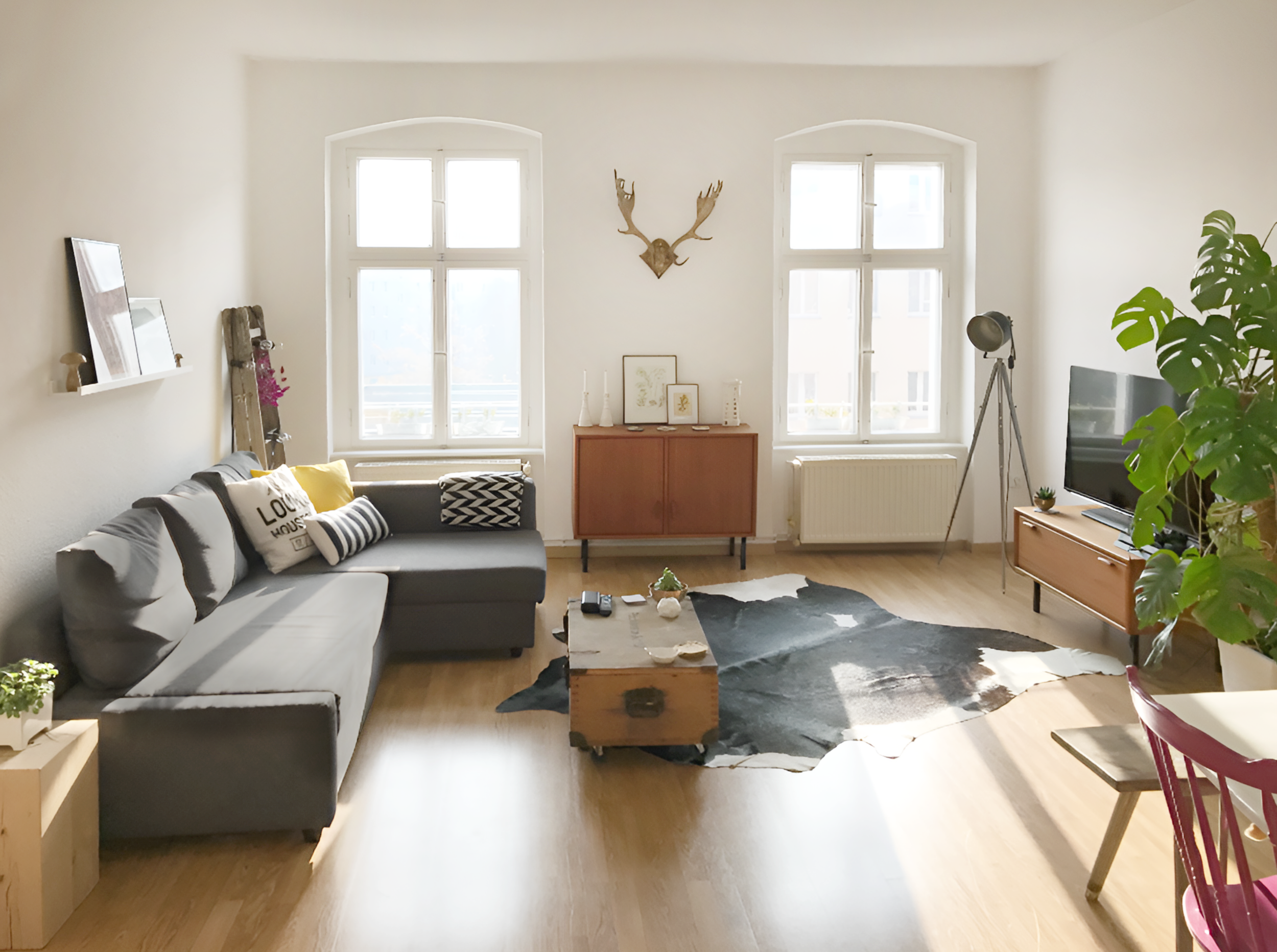 Sala de estar con sillón gris, mesa de centro de madera, alfombra en forma irregular, televisión en mueble de madera, ventana grande, piso de madera clara, plantas y decoración moderna.