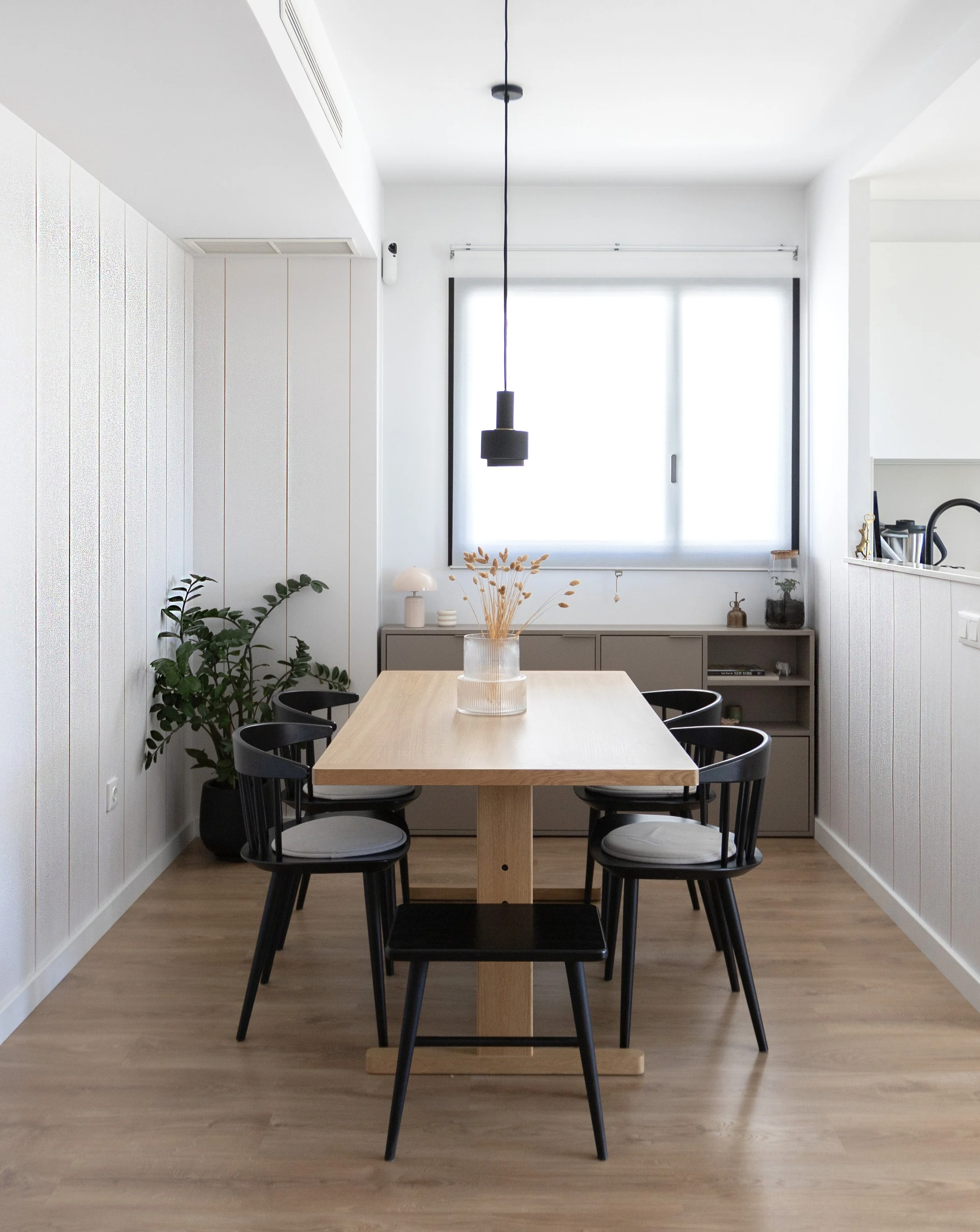 Comedor con mesa de madera y seis sillas negras, en una habitación luminosa con ventana grande, plantas decorativas y estanterías blancas.