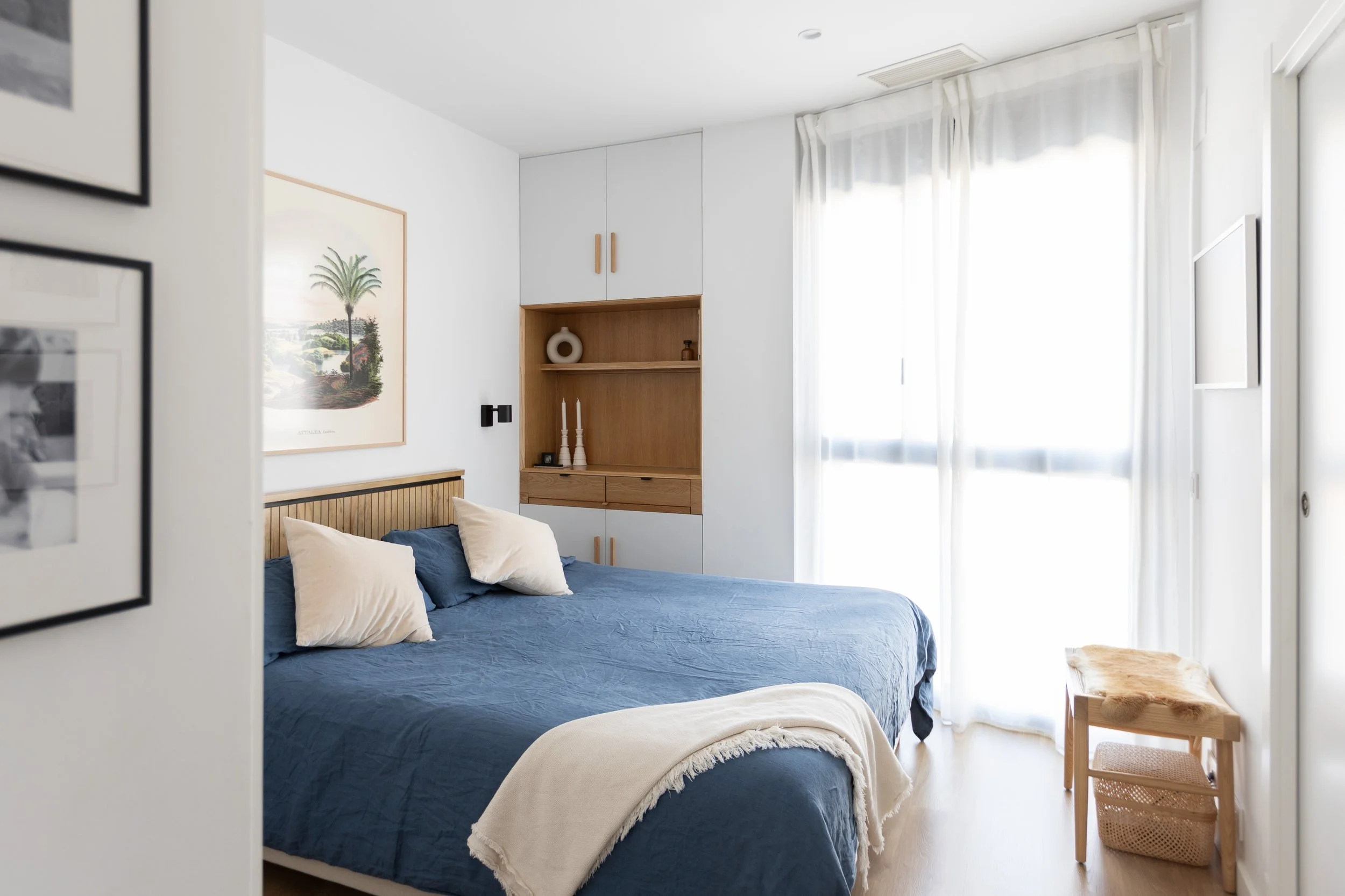 Dormitorio con cama de colchón azul, paredes blancas, ventanas con cortinas blancas y mueble de madera con decoración en tonos neutros.