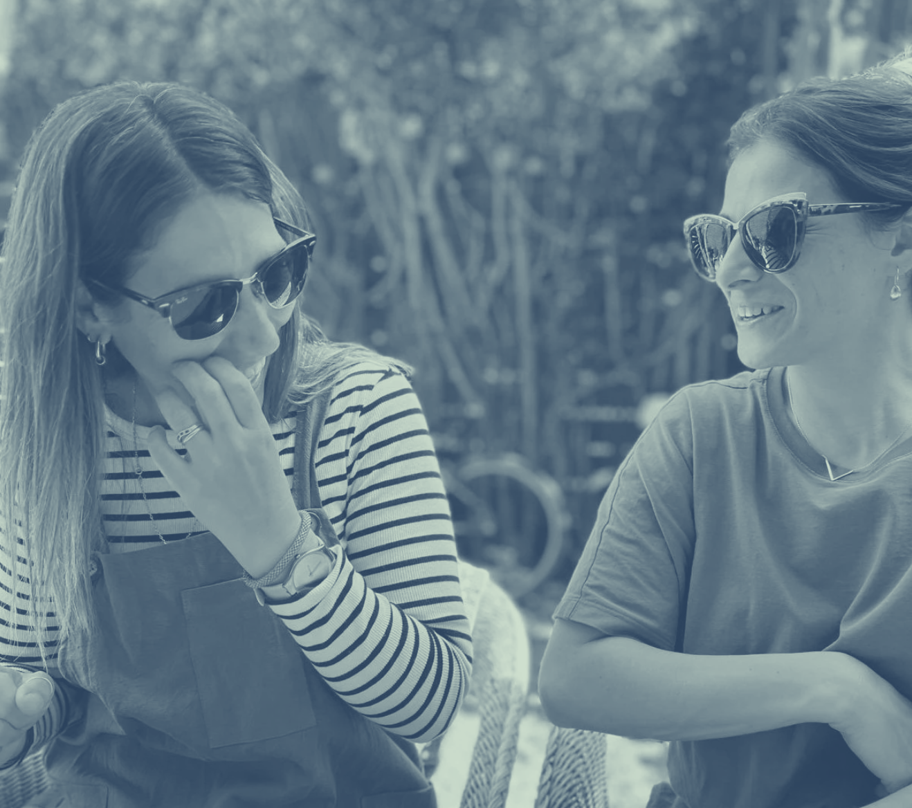 Dos mujeres con gafas de sol conversando al aire libre, en un entorno de jardín o parque.