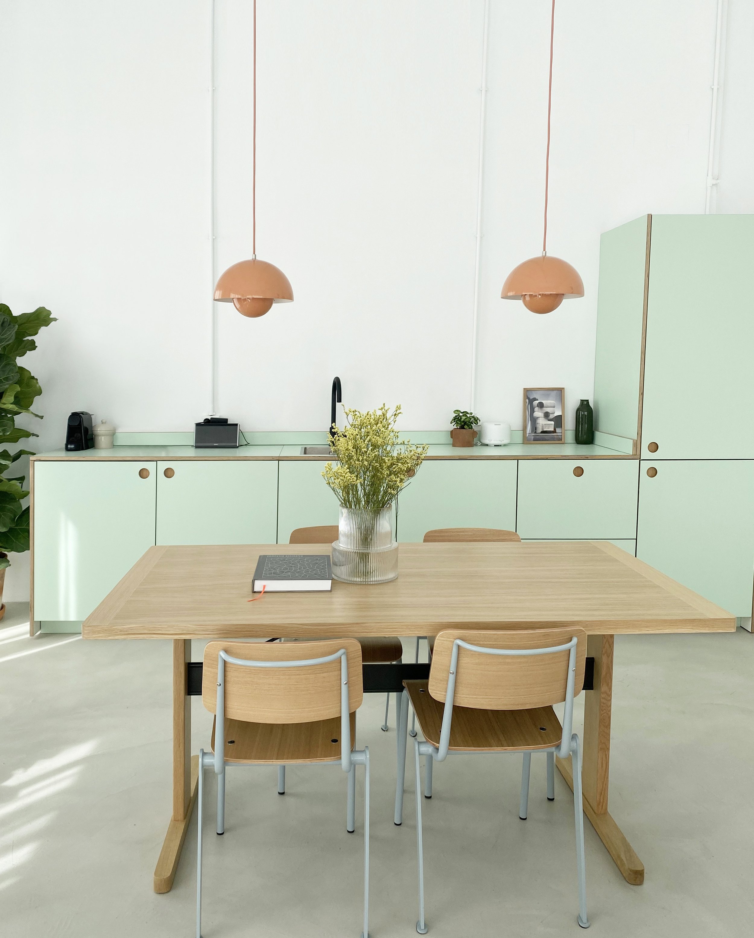 Una mesa de madera con cuatro sillas, un libro y un jarrón con flores. Al fondo, una cocina moderna con gabinetes mint y accesorios, dos lámparas de techo de color terracota, una planta, y algunos objetos decorativos.