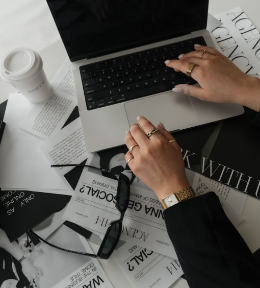 A person's hands working on a silver laptop surrounded by scattered papers, glasses, and a coffee cup on a desk.