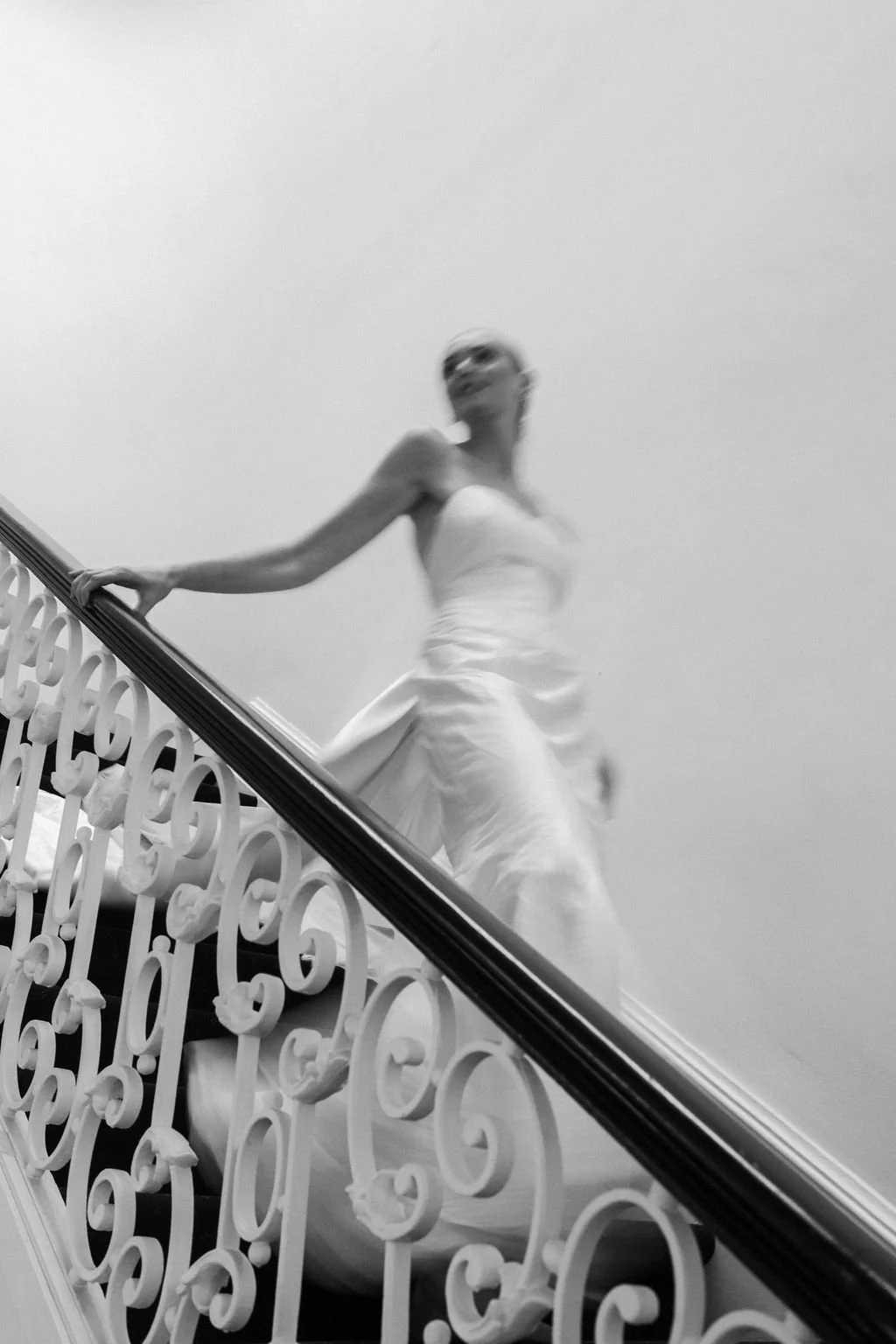 A woman in a white dress walking up a staircase with ornate railing, viewed from below, in black and white.