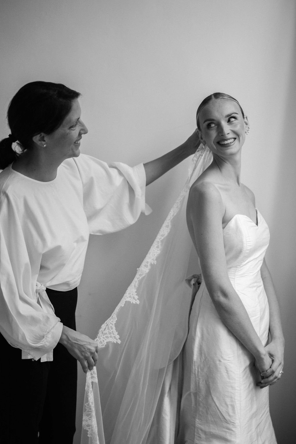 A smiling bride in a strapless wedding gown, looking to her right while a woman adjusts her veil.