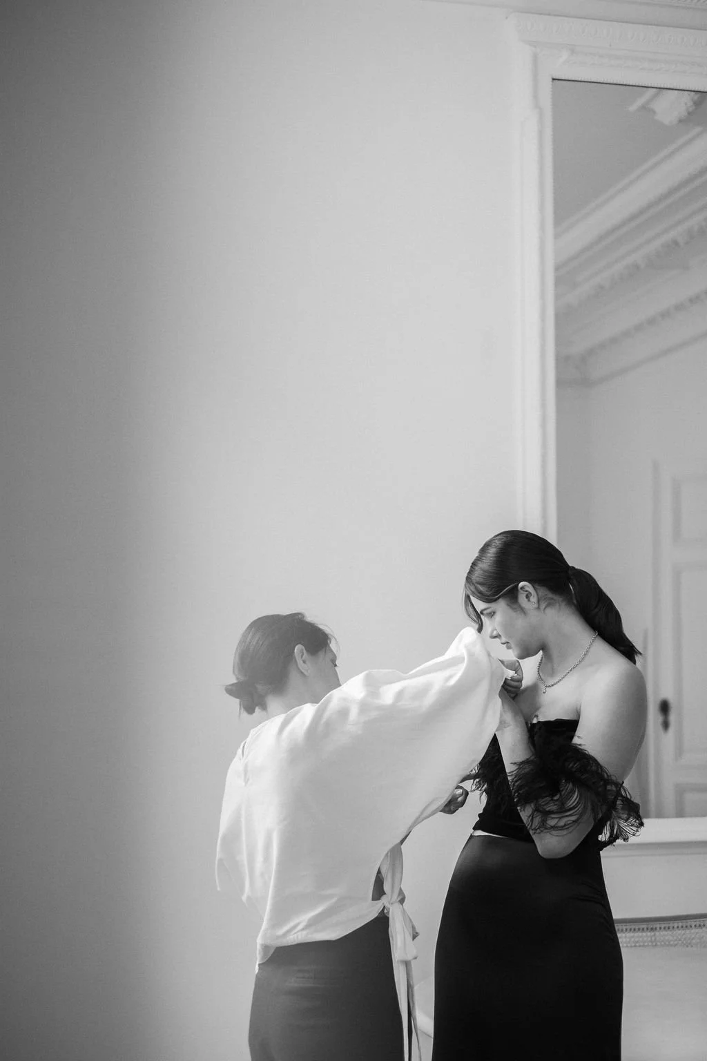A woman in a black dress with shoulder feathers is having her dress adjusted by another woman wearing a white top in a room with ornate molding and a large mirror.