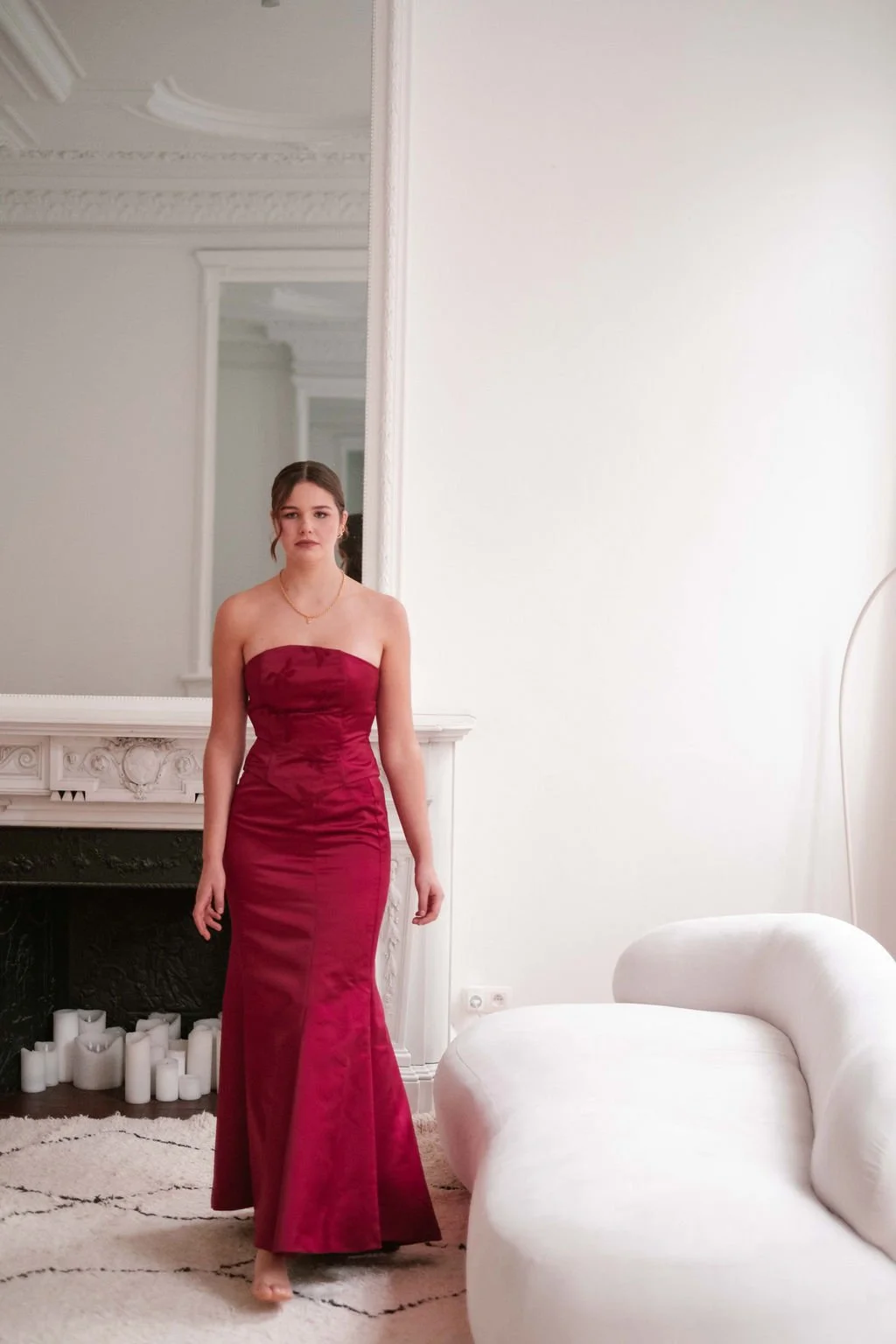 A young woman in a strapless red gown standing in a bright, decorated room with a mirror, white fireplace, candles, and a white couch.