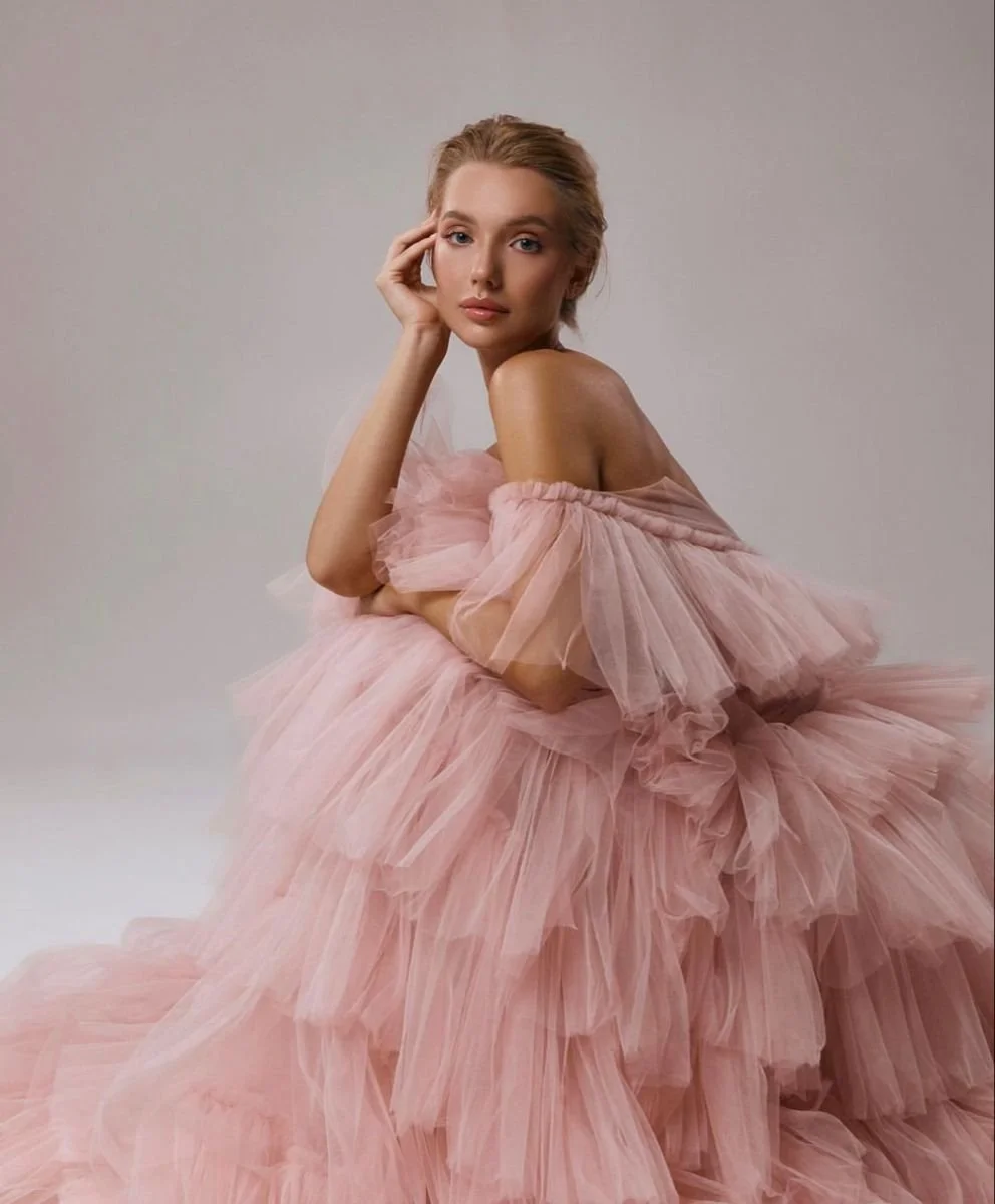 A woman wearing a flowing pink tulle dress with off-shoulder ruffles, sitting against a plain neutral background.