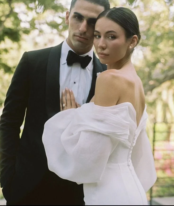 A man in a tuxedo and woman in a white off-shoulder dress pose closely outdoors, with trees in the background.