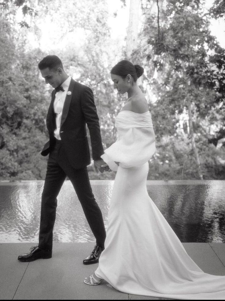 Black-and-white photo of a bride and groom holding hands and walking, outdoors near a water feature with trees in the background. The groom wears a tuxedo, and the bride wears a long, off-the-shoulder wedding gown.