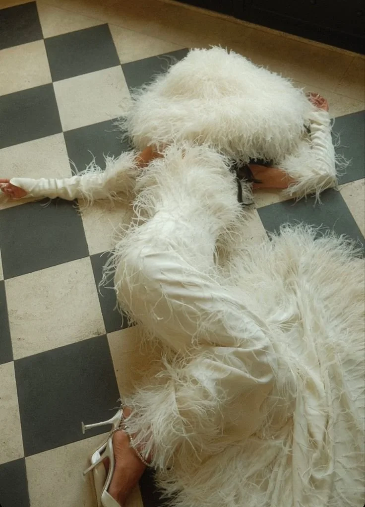 A person dressed in a long, white feathered dress lying face down on a black-and-white checkered floor, with their arms stretched out and wearing white high-heeled shoes.