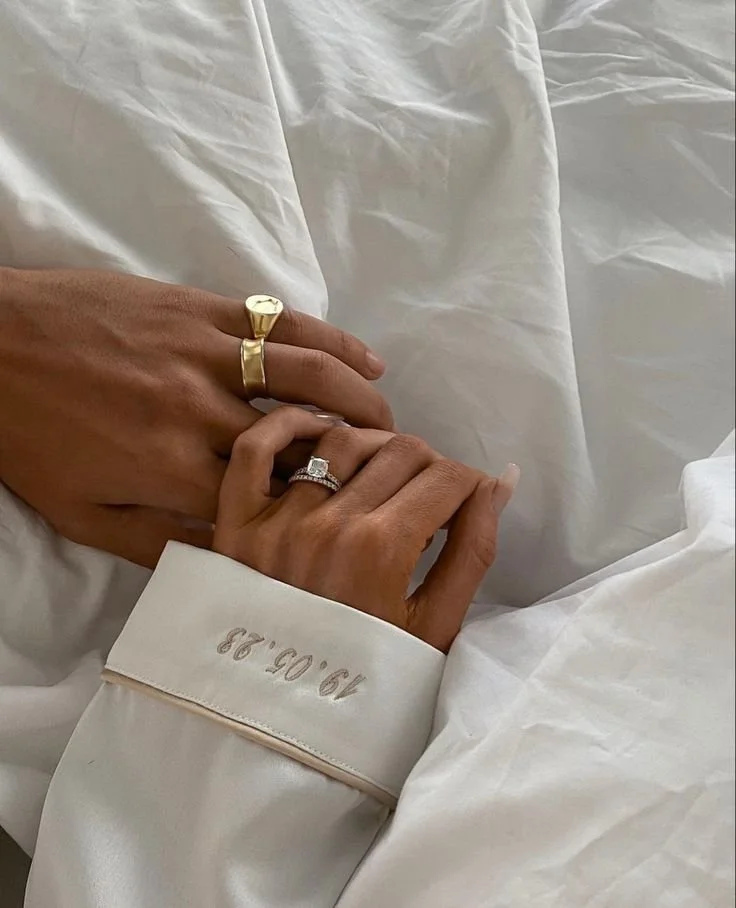Two hands with wedding rings, resting on white bedsheets. One hand has a gold ring and the other has a diamond ring. A white robe with a date embroidered on the sleeve is also visible.