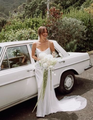 A woman in a white wedding dress holding a bouquet of white flowers, standing next to a vintage white car on a scenic outdoor road with greenery and trees in the background.