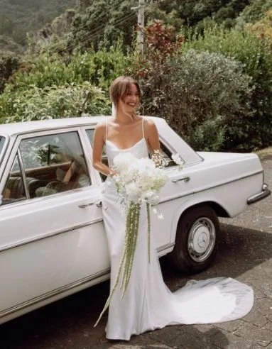 A woman in a white wedding dress holding a bouquet of white flowers, standing next to a vintage white car outdoors with trees and greenery in the background.