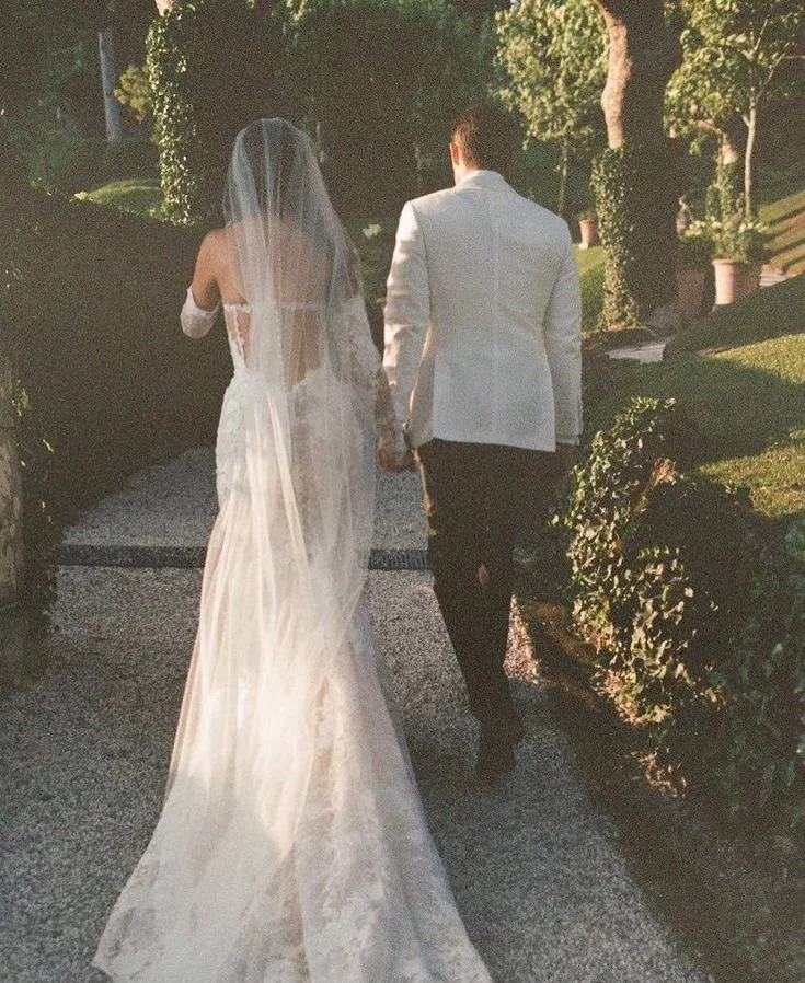 A bride in a white wedding dress and veil walking hand-in-hand with a groom in a white tuxedo jacket and black pants, outdoors on a sunny day.