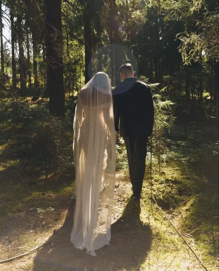 Couple dressed in wedding attire walking through a sunlit forest, with the bride in a white dress and veil and the groom in a dark suit.