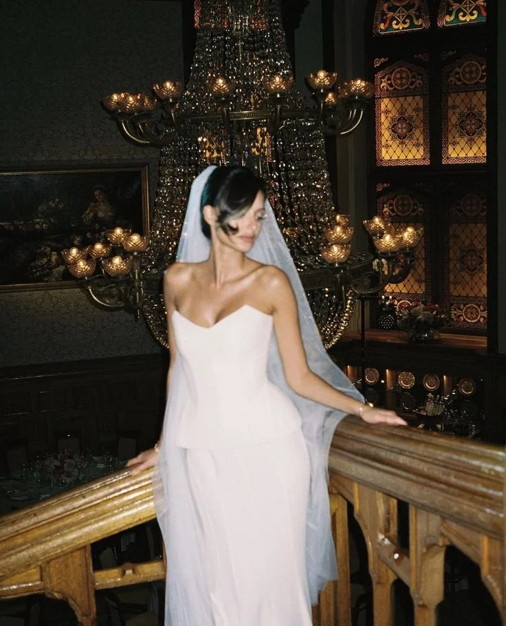 Woman in white wedding gown and veil standing on wooden staircase in an ornate, dimly-lit room with a large chandelier and stained glass windows.