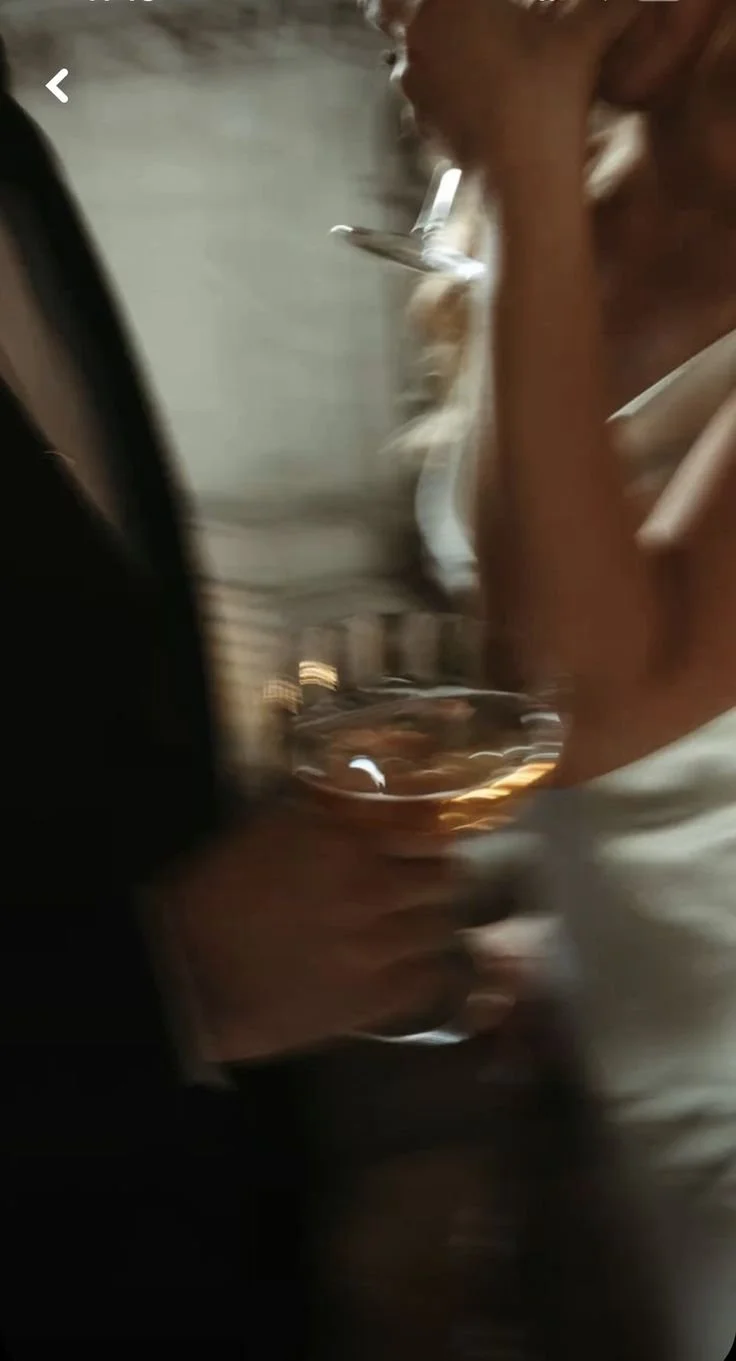 A woman with reddish hair drinking from a glass of brown-colored liquid, possibly whiskey or bourbon, with a slightly blurry background.