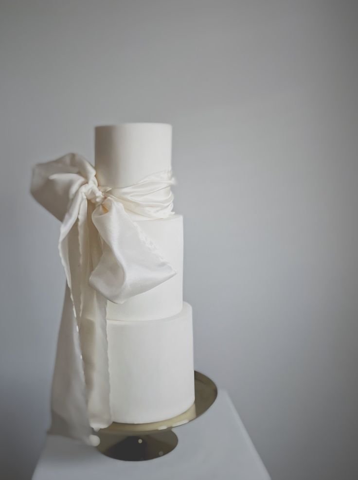 White tiered wedding cake with a large white ribbon bow on the side, placed on a white surface against a plain background.
