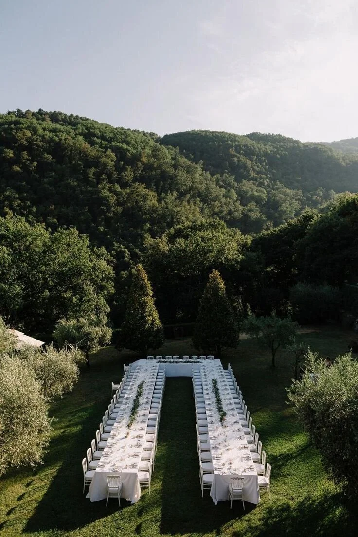 An outdoor dining setup with long white tables and chairs, arranged in a U-shape on a grassy area, surrounded by trees with forested mountains in the background.
