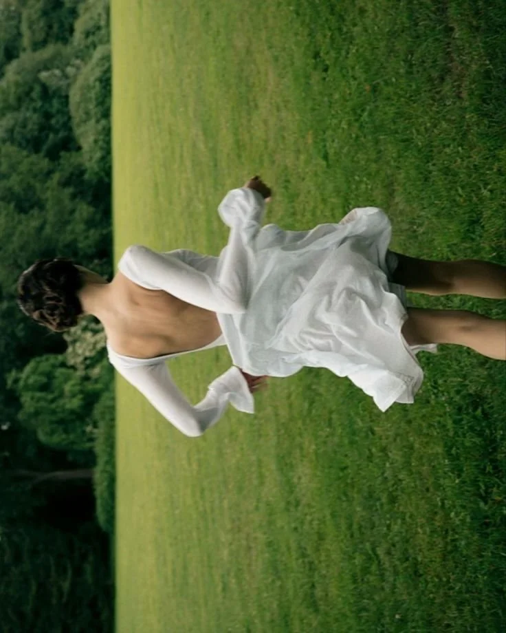 A woman walking outdoors on a grassy field, with her back turned, wearing a white dress and a white cardigan draped over her shoulders, surrounded by trees and greenery.