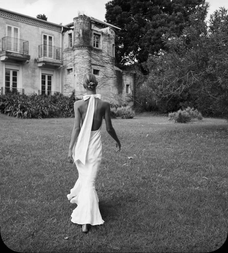 A woman walking away from the camera in a flowing dress with a bow around her neck on a lawn, with an old house and trees in the background, in black and white.