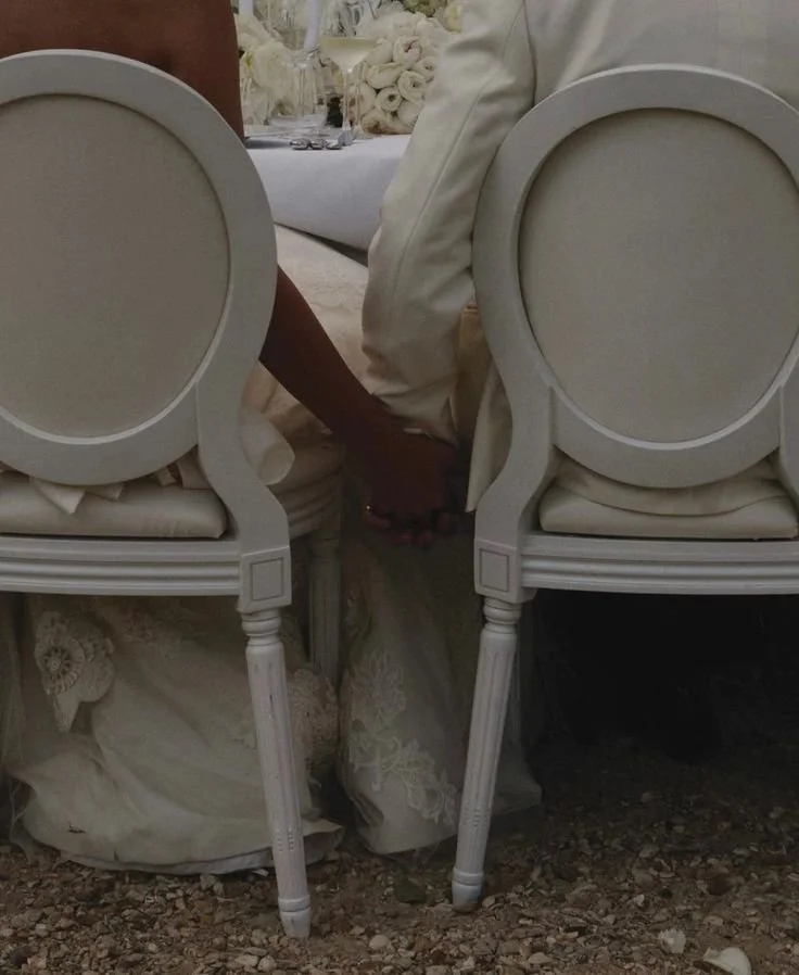 A bride and groom holding hands, sitting at a wedding reception table with white chairs and floral decorations.