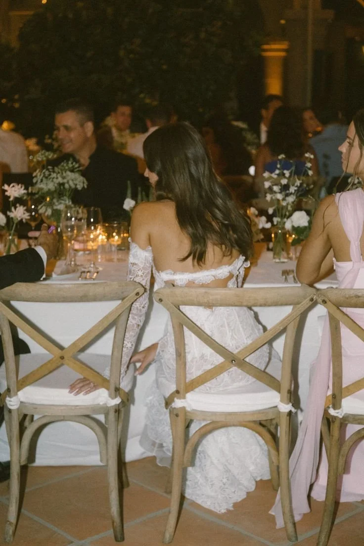 A woman in a white lace dress seated at an outdoor dinner table during a nighttime event with other guests and floral centerpieces.