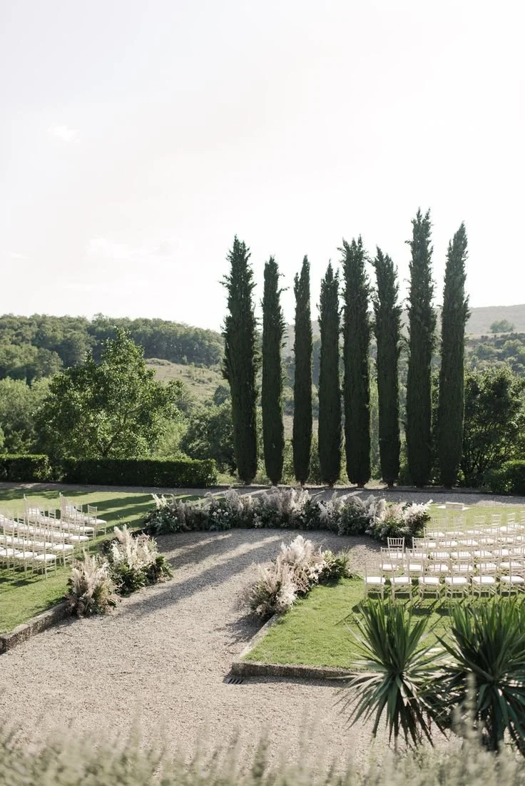 Outdoor wedding ceremony setup with white chairs arranged on either side of a gravel aisle, decorated with floral arrangements, in front of tall cypress trees and green rolling hills.