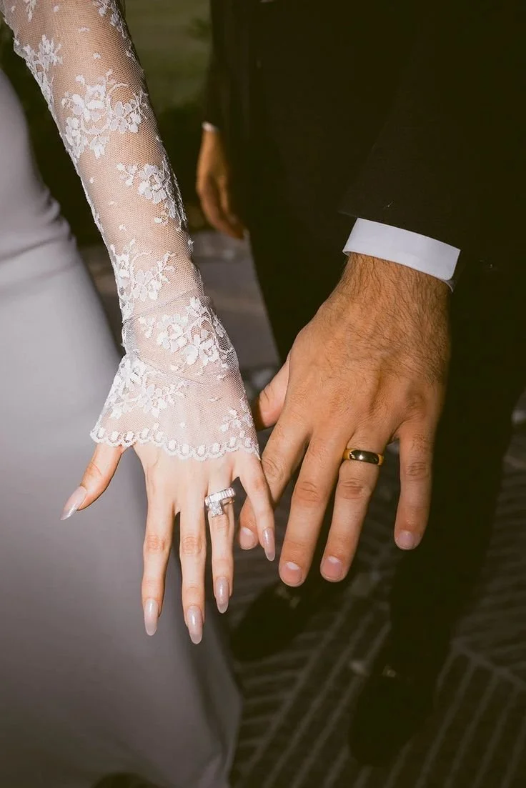 Close-up of a bride and groom's hands showing their wedding rings, with the bride wearing a lace long-sleeve dress and the groom in a black suit.